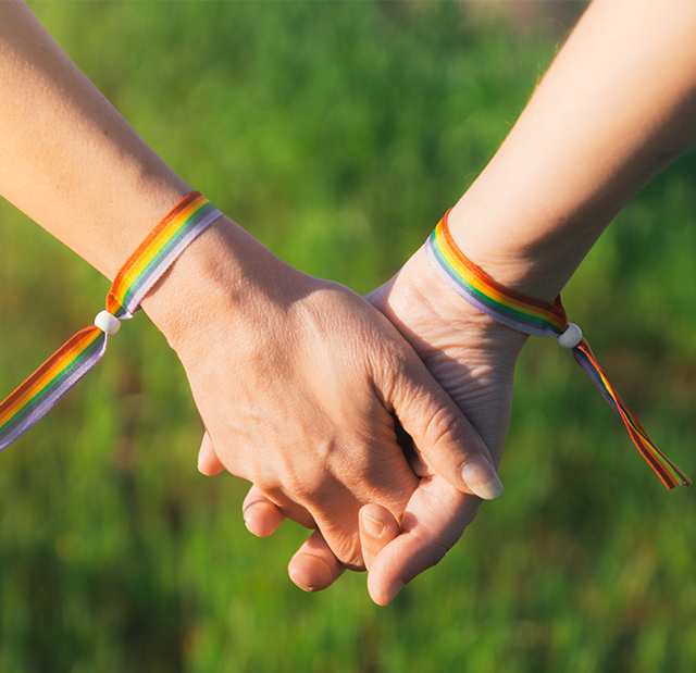 An up close shot of two people holding hands, who are both wearing rainbow pride festival bracelets