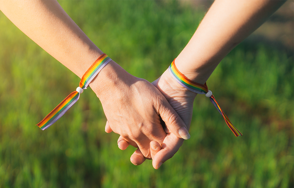 An up close shot of two people holding hands, who are both wearing rainbow pride festival bracelets